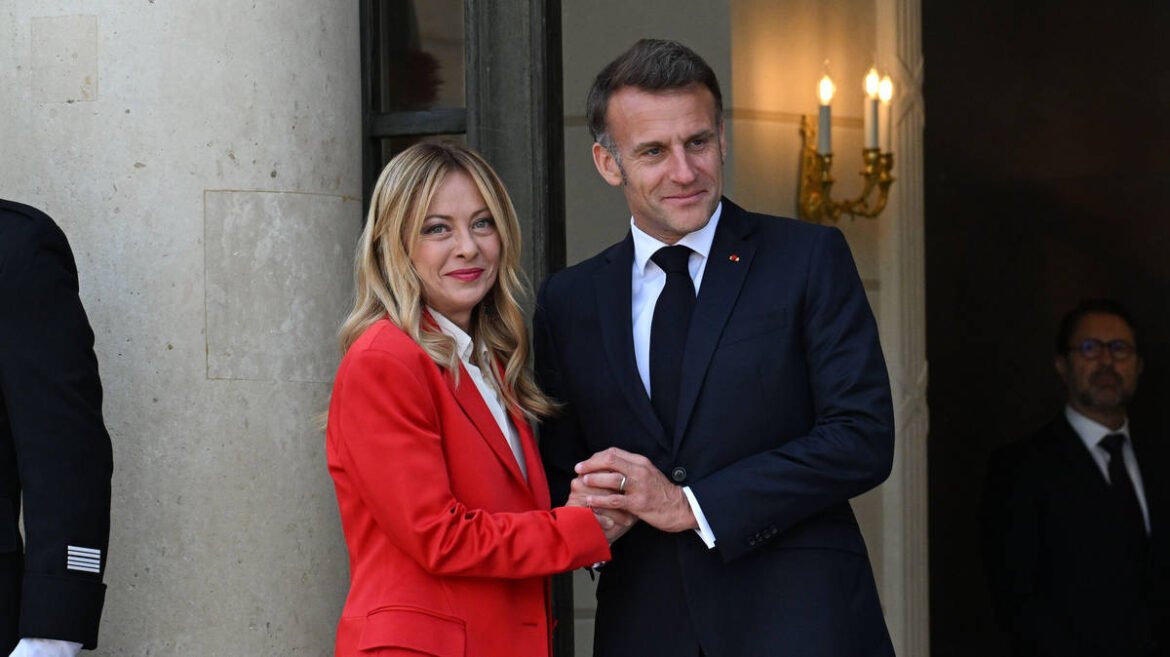 A man in a dark suit and tie and a woman in a red blazer shake hands outside a stone building, smiling at the camera.