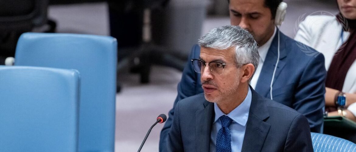 Man with gray hair and glasses in a navy suit speaks into a microphone at a formal meeting setting.