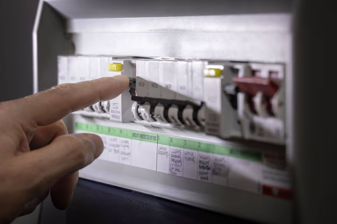 Person's finger presses a switch on an electrical distribution panel with labeled breakers and wiring visible in the background.