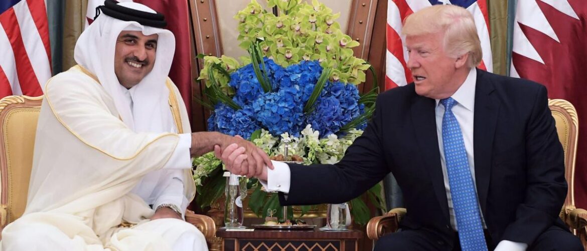 Two men in formal attire shake hands in front of a floral centerpiece and U.S. flags during a meeting.