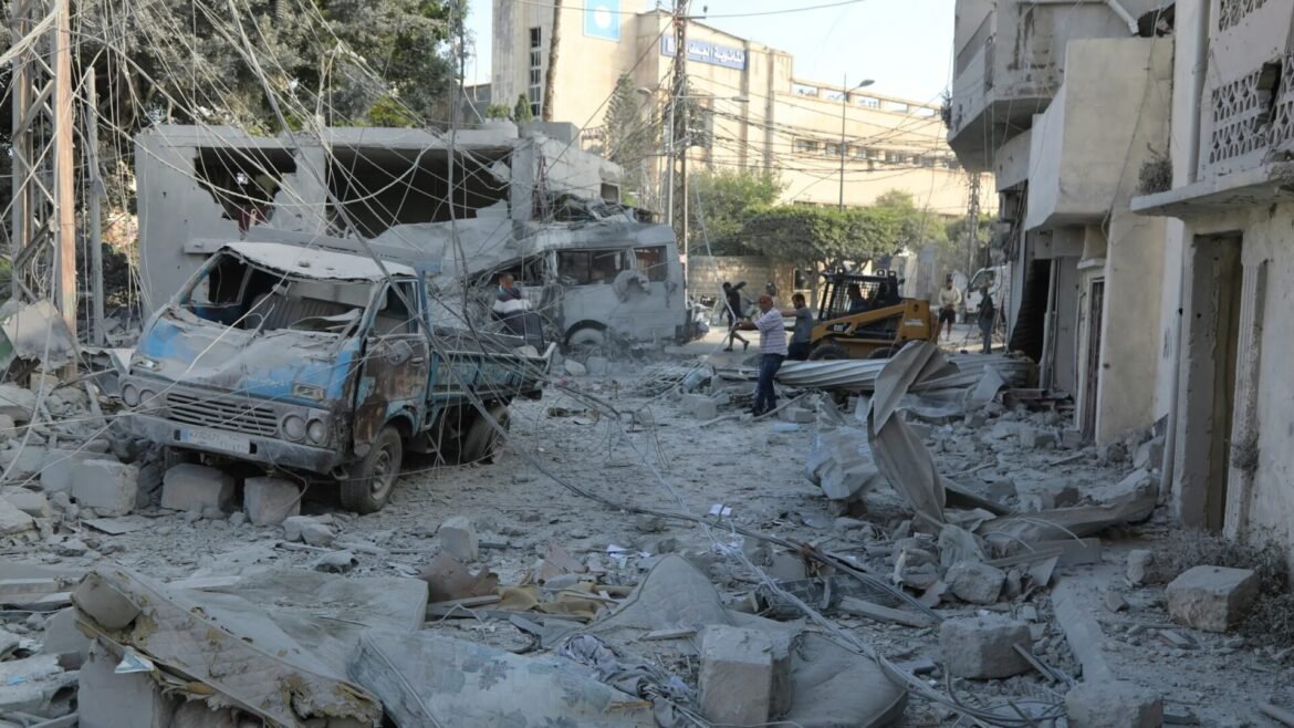War-ravaged street with collapsed buildings, tangled wires, and a damaged blue bus buried in rubble; workers survey the destruction and a skid-steer loader is in the background.
