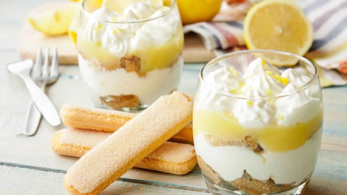 Two lemon dessert parfaits in clear glasses with whipped cream and biscuit layers, lemon slices in the background.