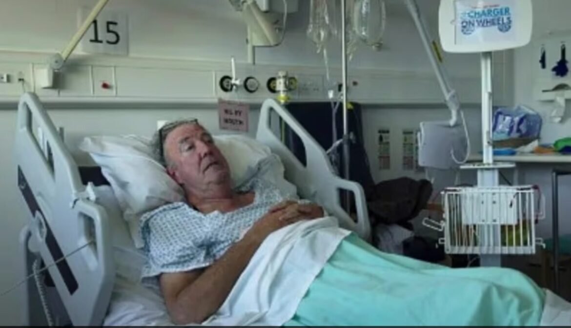 Elderly man lying in a hospital bed, awake with hands on chest, medical equipment and monitors around the room.