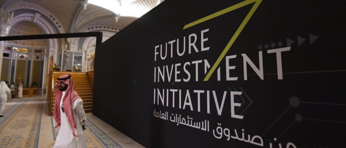 Person wearing traditional white thobe and red‑checkered ghutra walks past a large black banner that reads 'Future Investment Initiative' in a grand indoor hall.