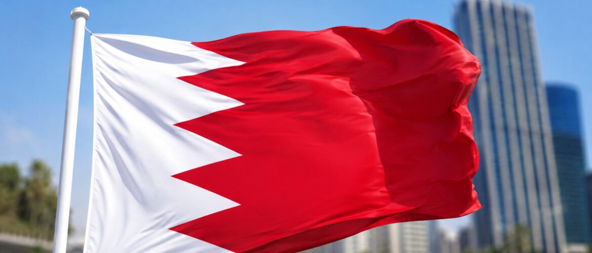 Flag of Bahrain waving in front of a city skyline with skyscrapers in the background, blue sky overhead.