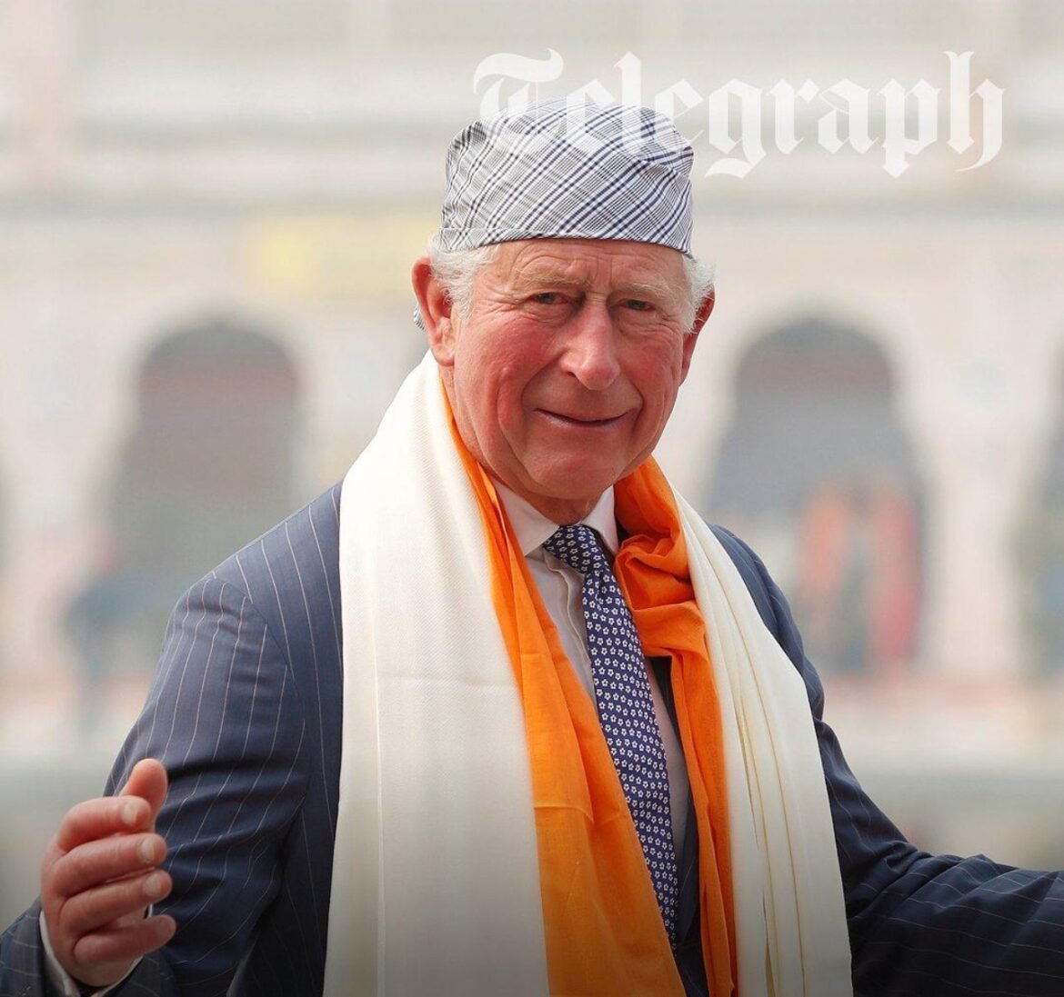 Older man in a pinstripe suit with orange and white scarves, wearing a patterned cap and smiling outdoors.