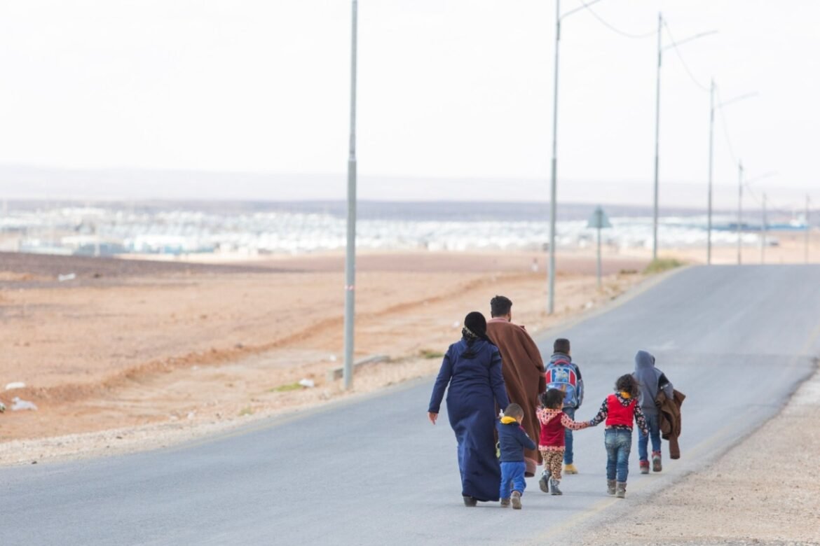 A family of six or more walking along a rural paved road in a barren desert landscape, holding hands, with utility poles lining the road and distant coastline on the horizon.