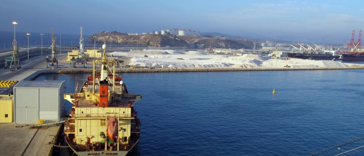 Large cargo ship docked at a busy harbor with cranes, warehouses, and a sandy storage area along the shoreline; calm blue water fills the foreground and distant hills rise beyond the port.