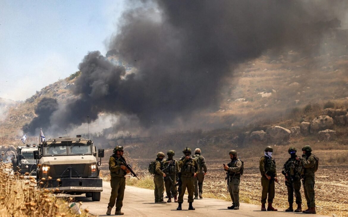 Soldiers in desert terrain stand near armored vehicles as thick black smoke rises from hills in the background.