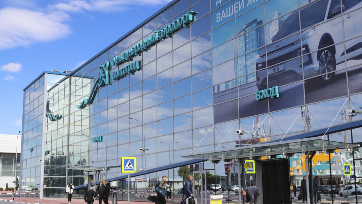 Glass-front international airport terminal with blue-tinted panels, Cyrillic signage, and people walking toward the entrance.