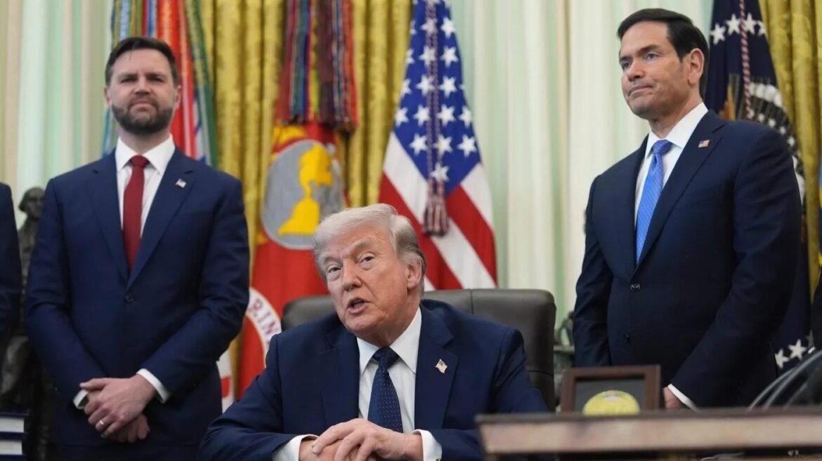 A man in a dark suit sits at a desk, flanked by two suited men standing, in a formal room with flags in the background.