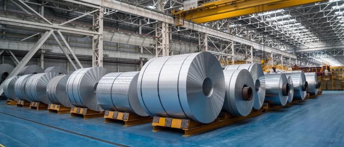 Row of large steel coils resting on yellow supports inside a spacious industrial warehouse.
