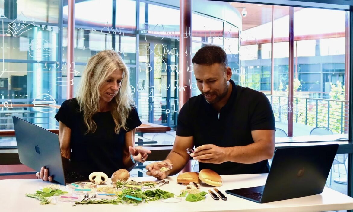 Two coworkers sit at a white table in a glass-walled office, examining food samples with open laptops nearby and herbs and bread on the table.