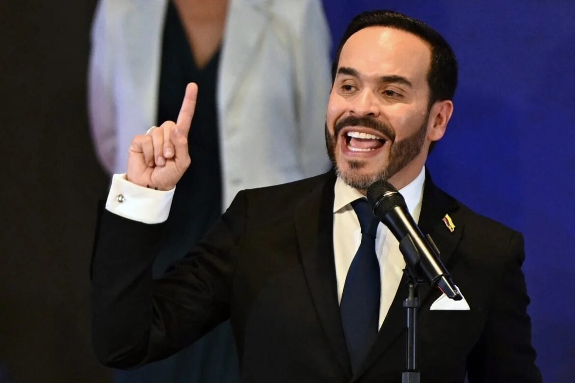 Man in a dark suit and blue tie speaks into a microphone, raising one finger in emphasis on stage with a blue backdrop.