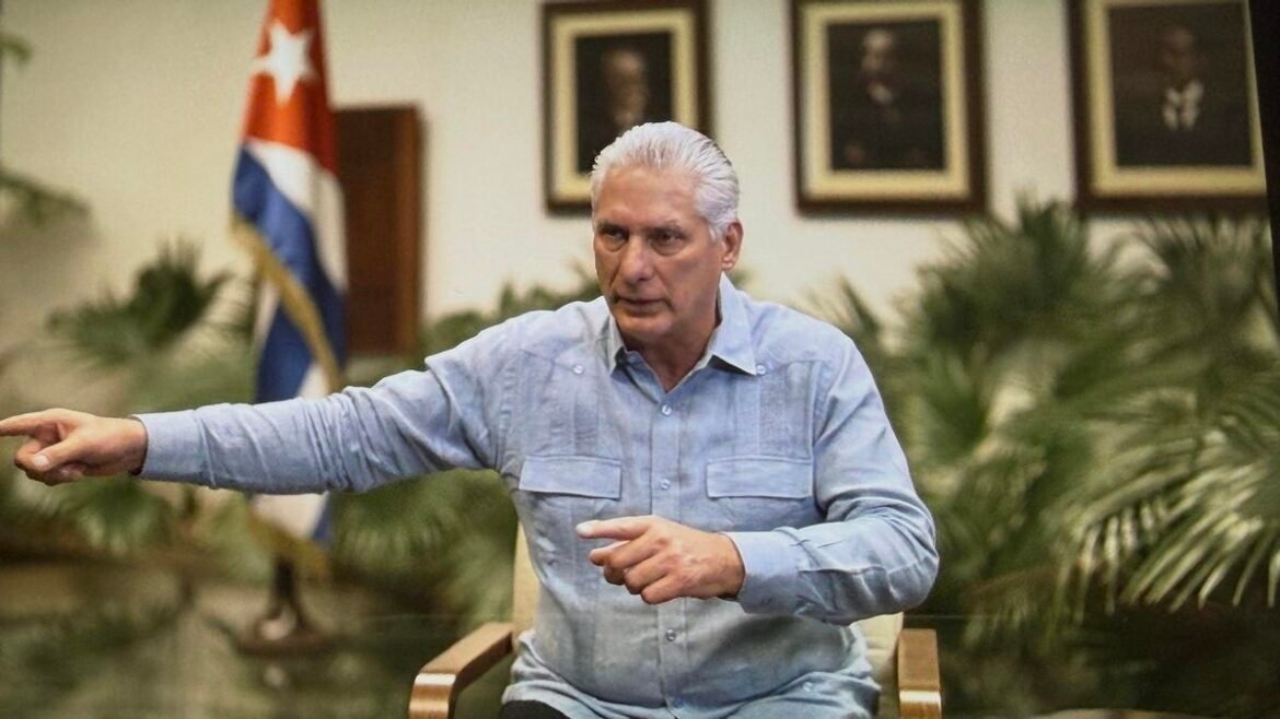 Older man with silver hair in a light blue shirt sits and points to the left in a formal room with a Cuban flag and framed portraits in the background.