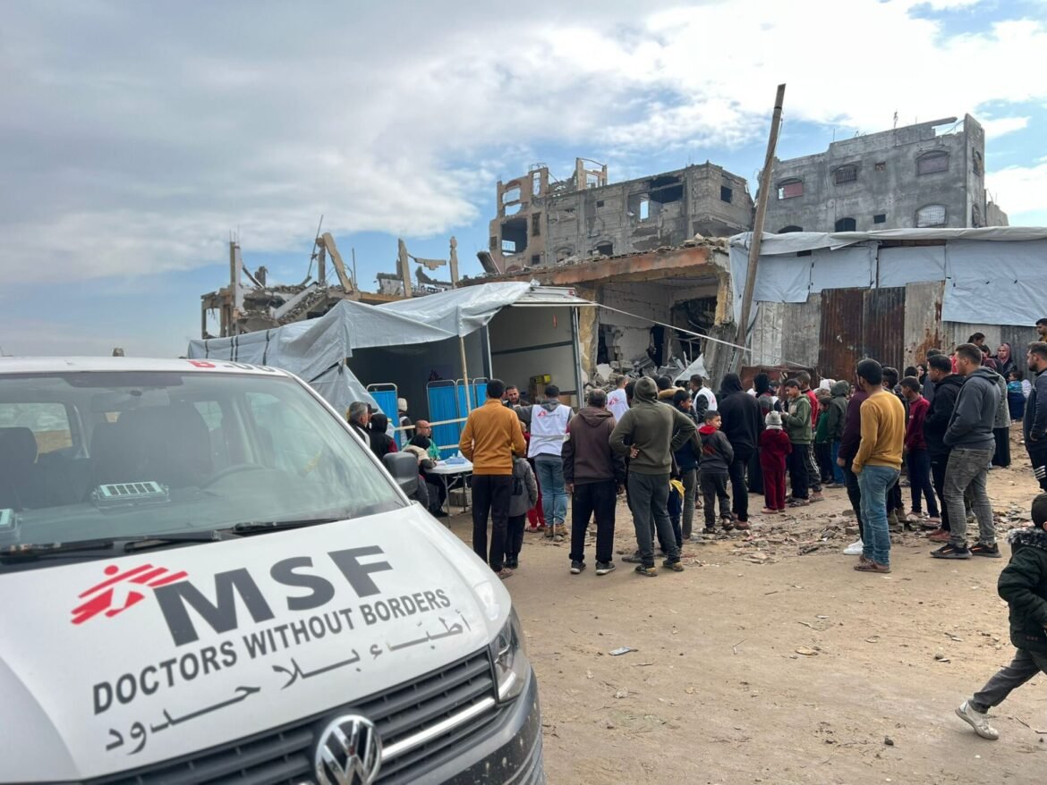 MSF van in foreground at a disaster site with a crowd gathered beside ruined buildings awaiting aid relief services