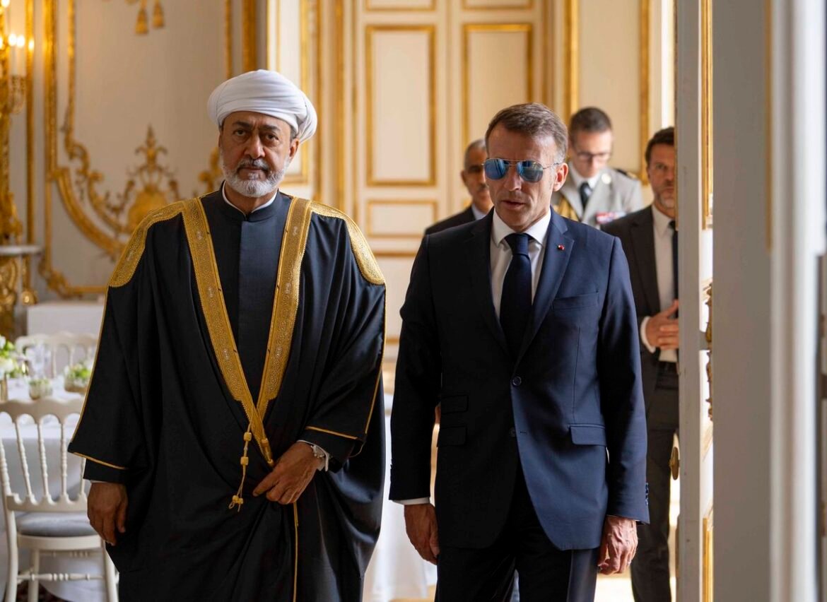 Two officials in formal dress walk through an opulent, gold-trimmed room; one in a black ceremonial robe with a white turban, the other in a navy suit and sunglasses.