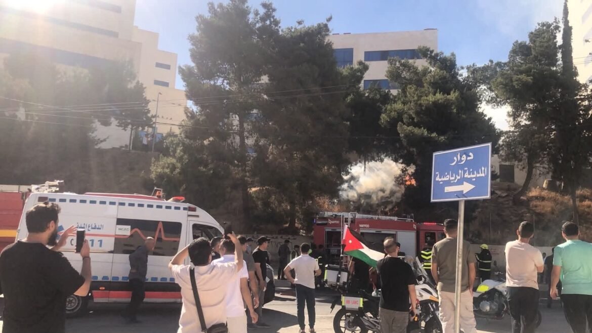 Emergency scene with an ambulance and a fire truck, bystanders filming and smoking visible behind trees under sunny skies, Arabic sign nearby.