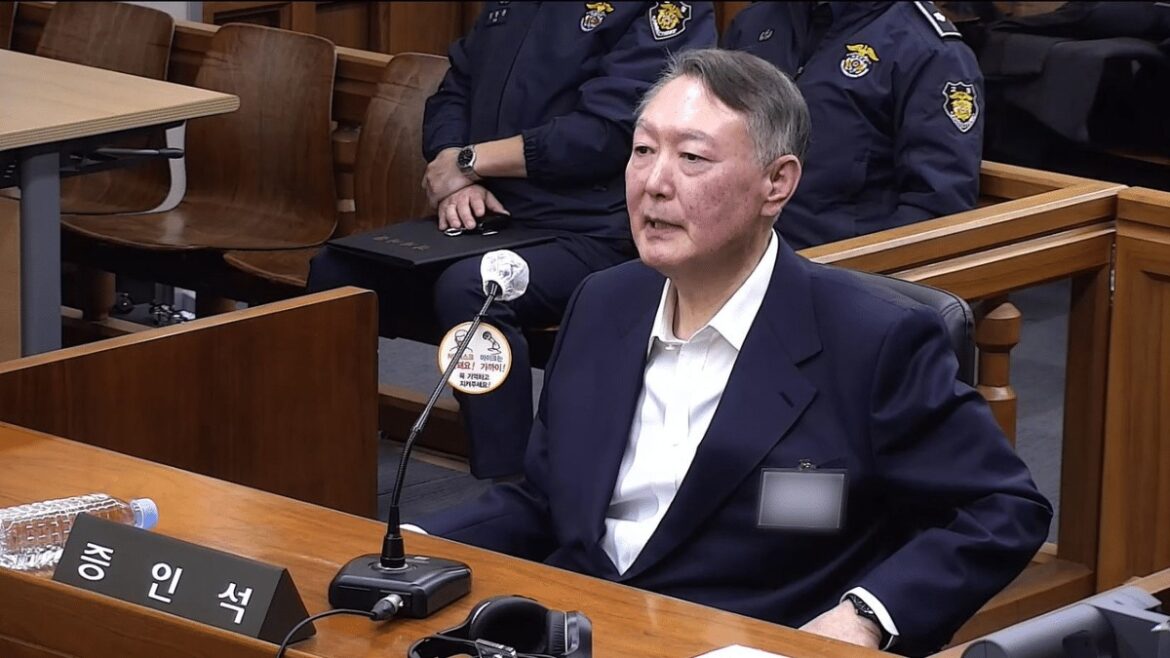 Man in a dark suit testifying at a hearing, seated at a wooden desk with a microphone in front of him in a courtroom setting with officers in the background.