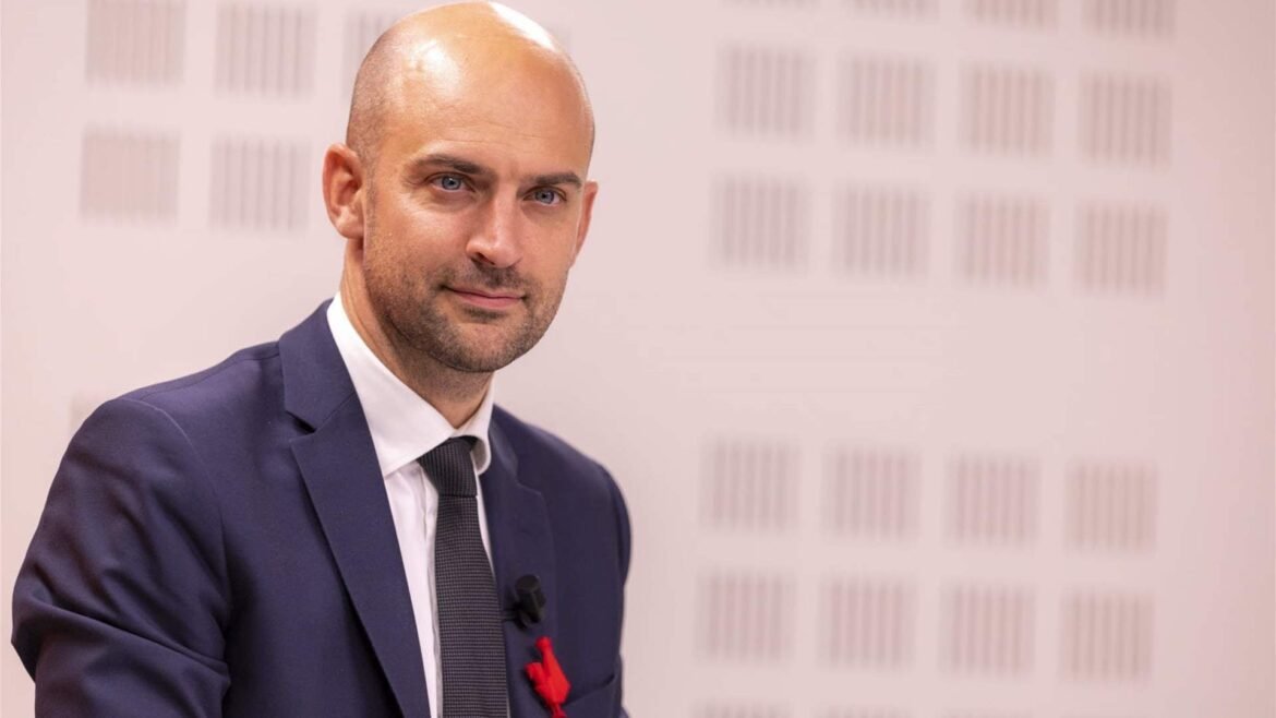 Portrait of a bald man in a navy suit and gray tie, sitting for an interview against a light patterned wall.