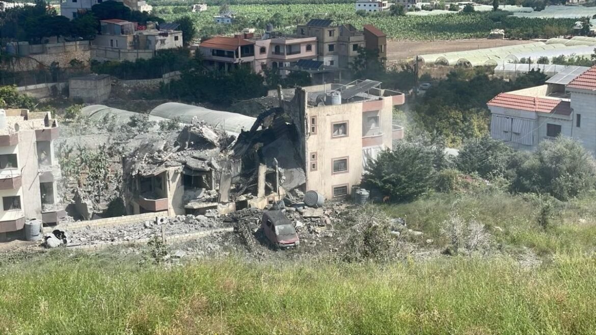 Rubble-filled scene of a collapsed multi-story house with debris and a red car among the rubble in a residential hillside area