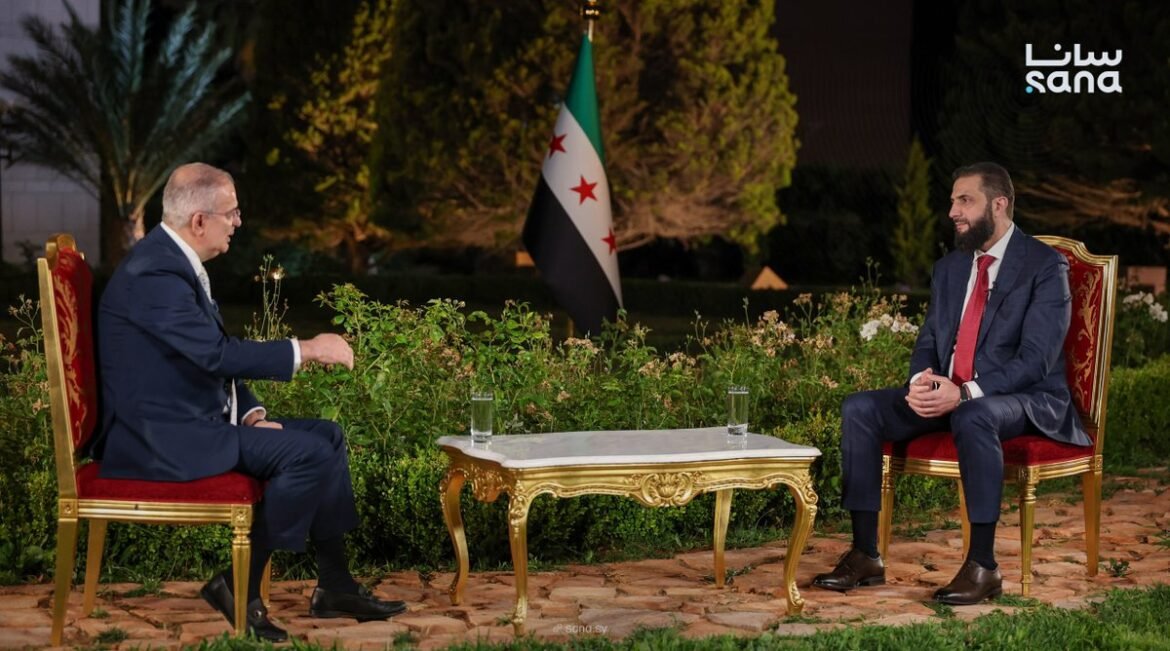 Two men in suits sit on ornate red chairs facing each other outdoors at night, with a marble table between them and a Syrian flag in the background.
