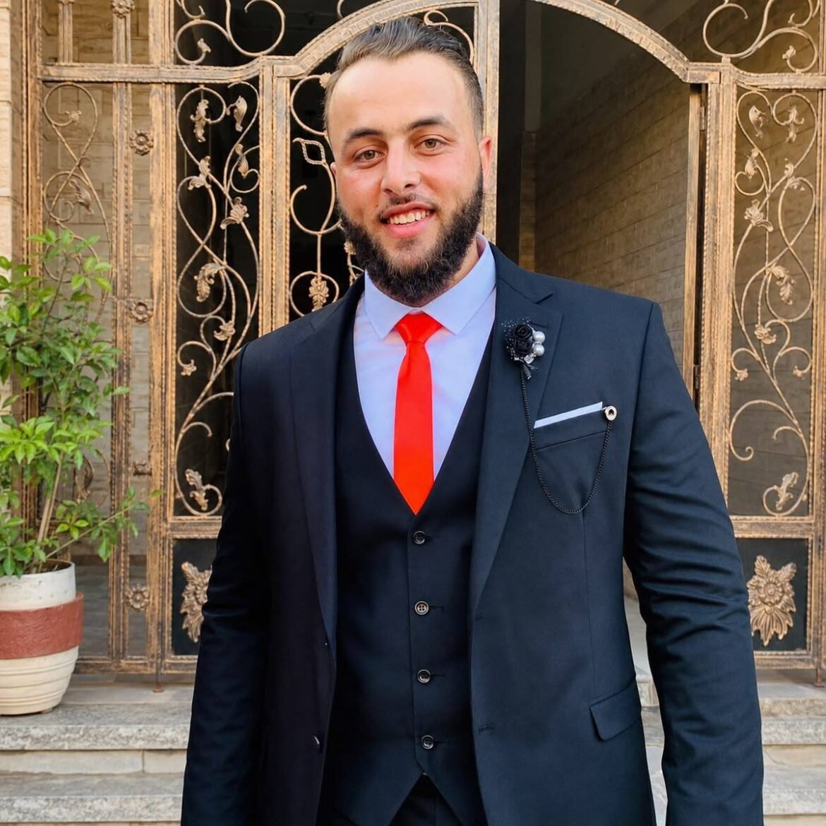Man in a dark suit with a red tie and matching vest, standing in front of an ornate gold gate outdoors.