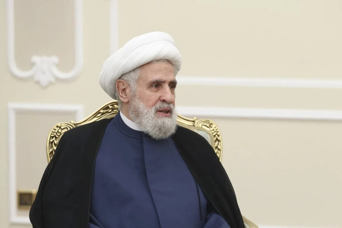 Man with a white turban and beard sitting on an ornate gold chair in a formal, beige-room setting; wearing dark blue robe and black cloak.