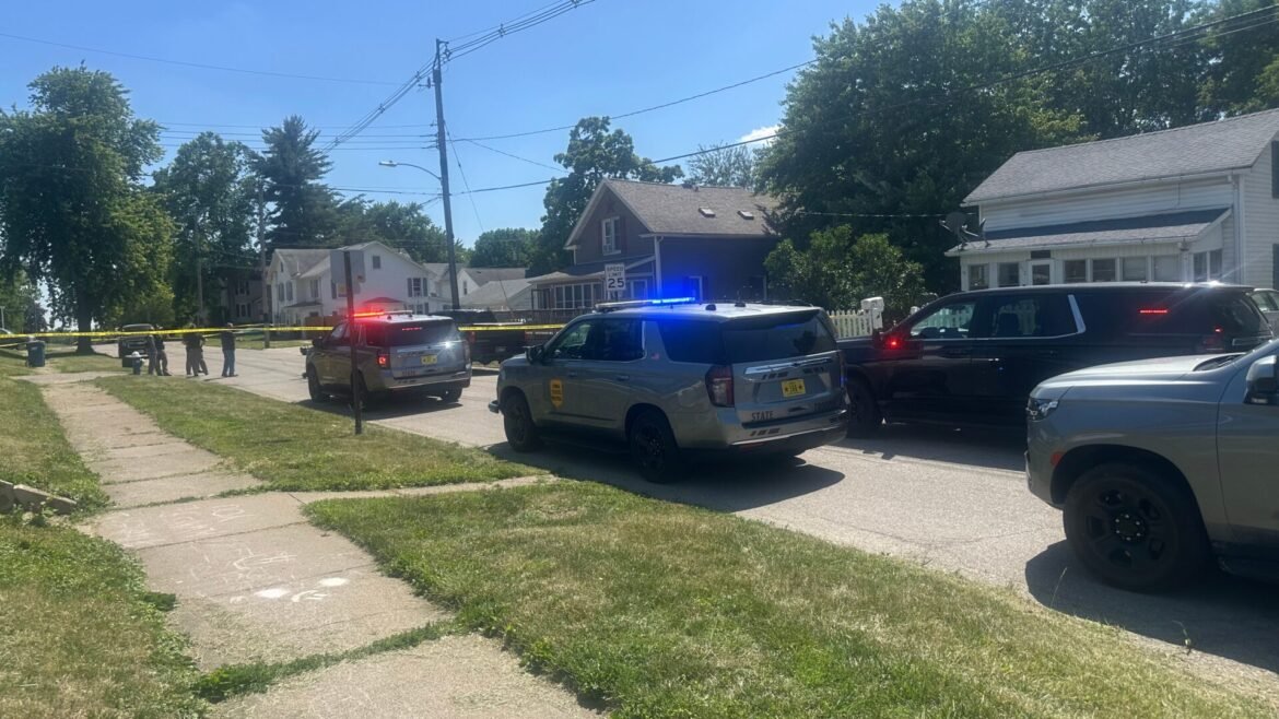 Police cars with flashing blue and red lights on a residential street, yellow caution tape cordoning off the area and people standing nearby.