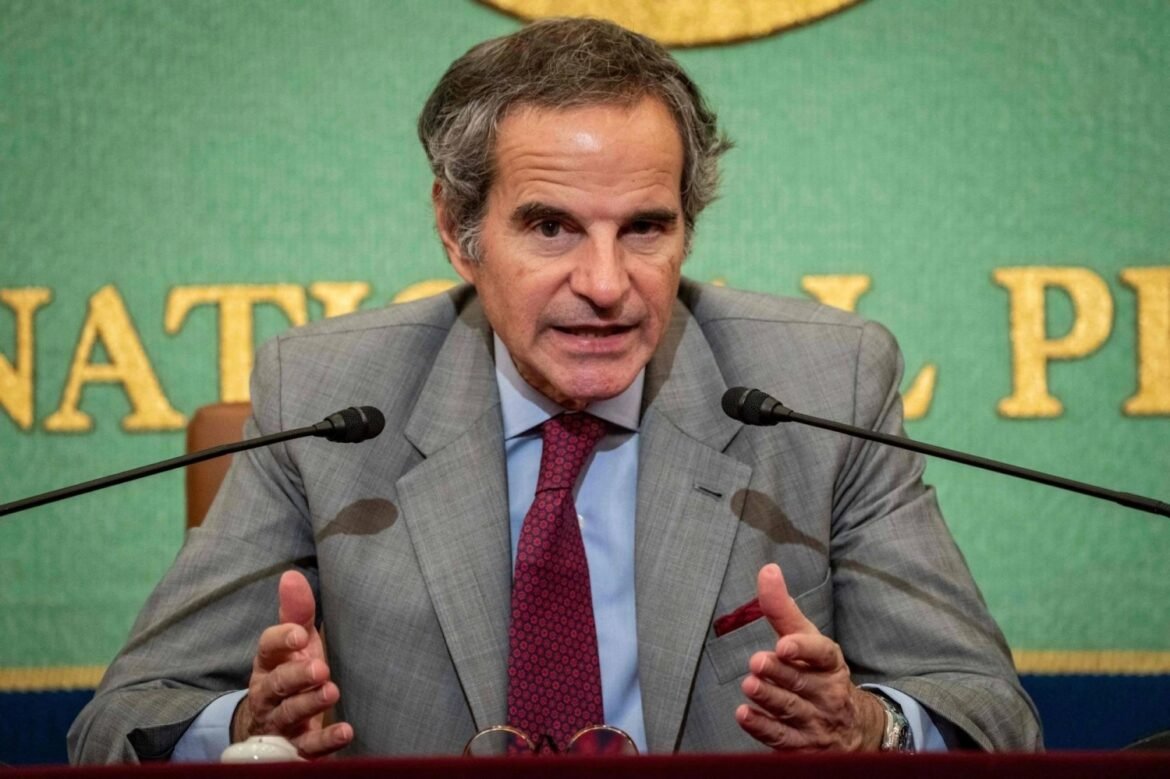 Middle-aged man in a gray suit and red tie speaks at a press conference, with two microphones in front and a green backdrop behind him.