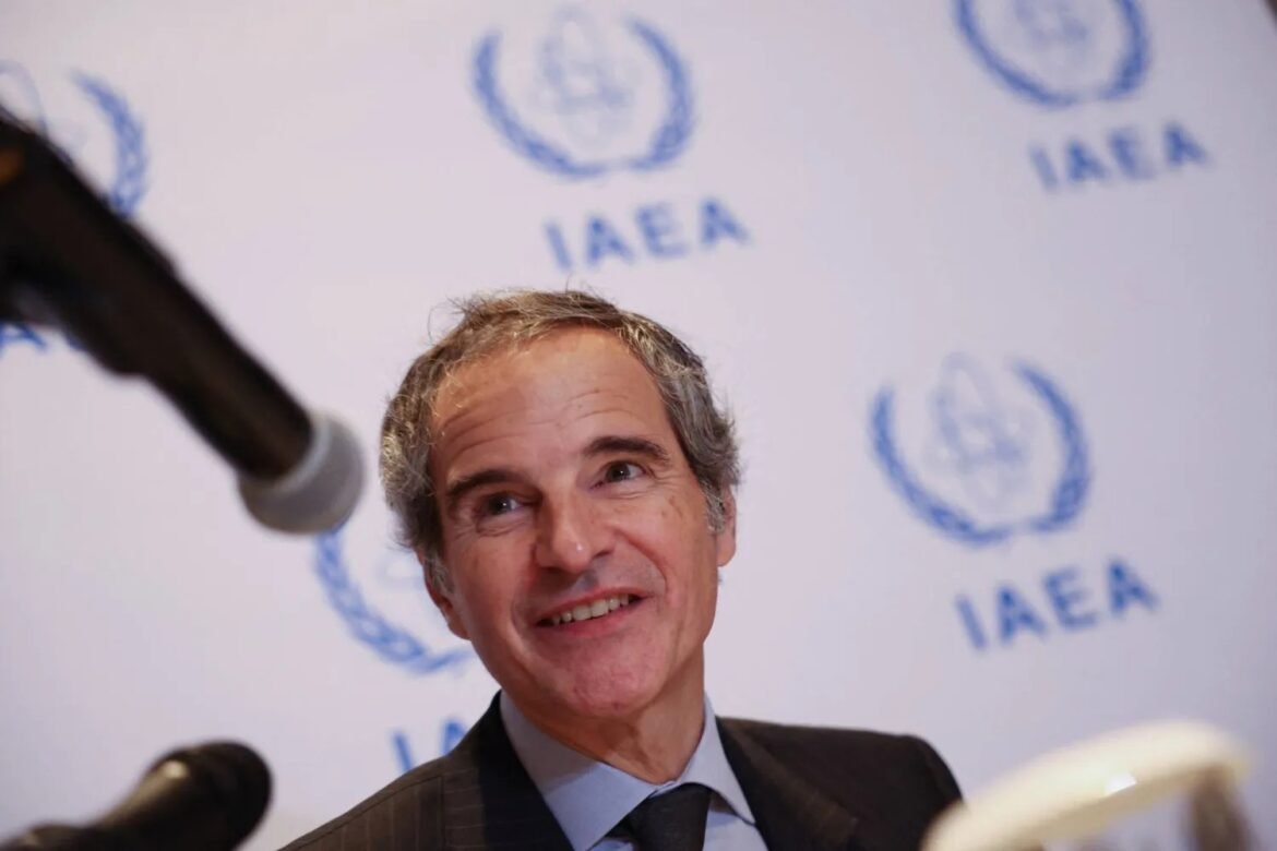 Smiling man in a dark suit and blue tie at a press conference, with an IAEA logo backdrop and microphones in the foreground.