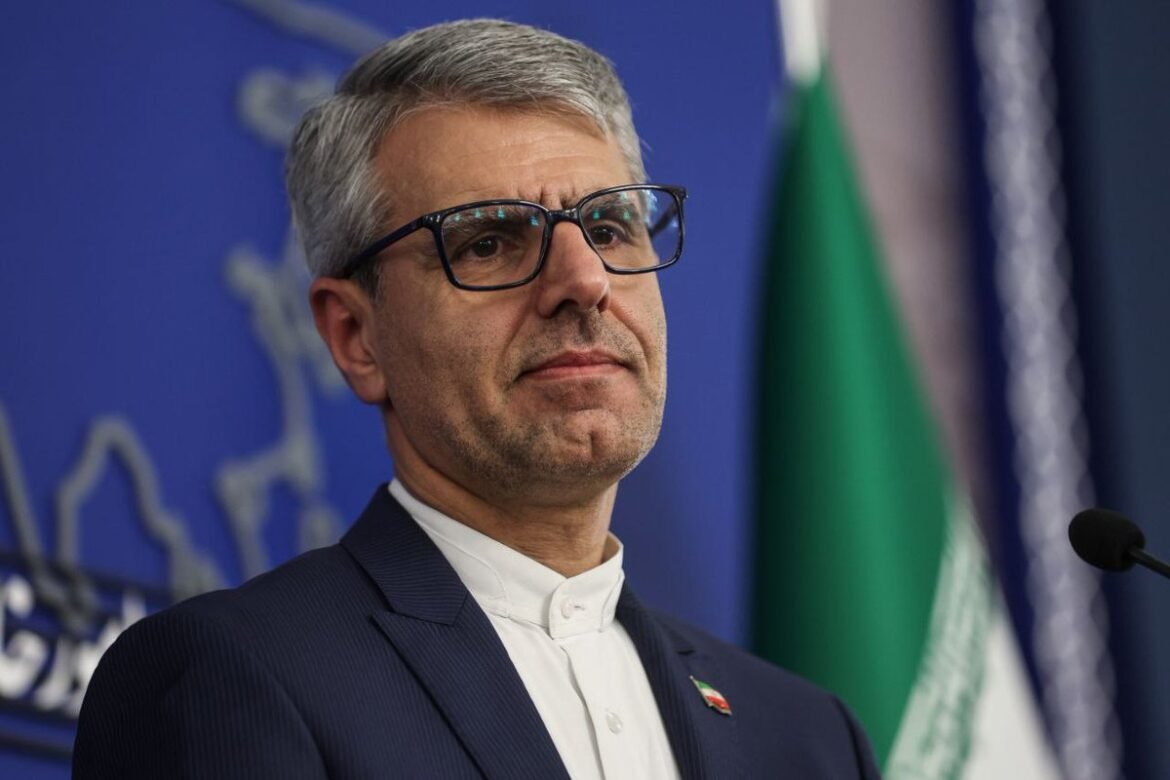 Man in a suit and glasses speaking at a press conference, with a blue backdrop and flags nearby.