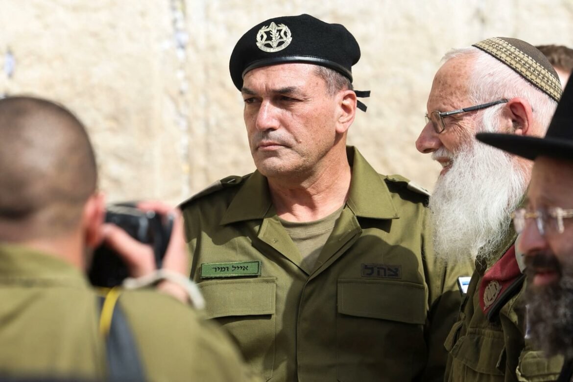 Military officer in olive uniform and black beret stands with other soldiers, a photographer in the foreground films him.