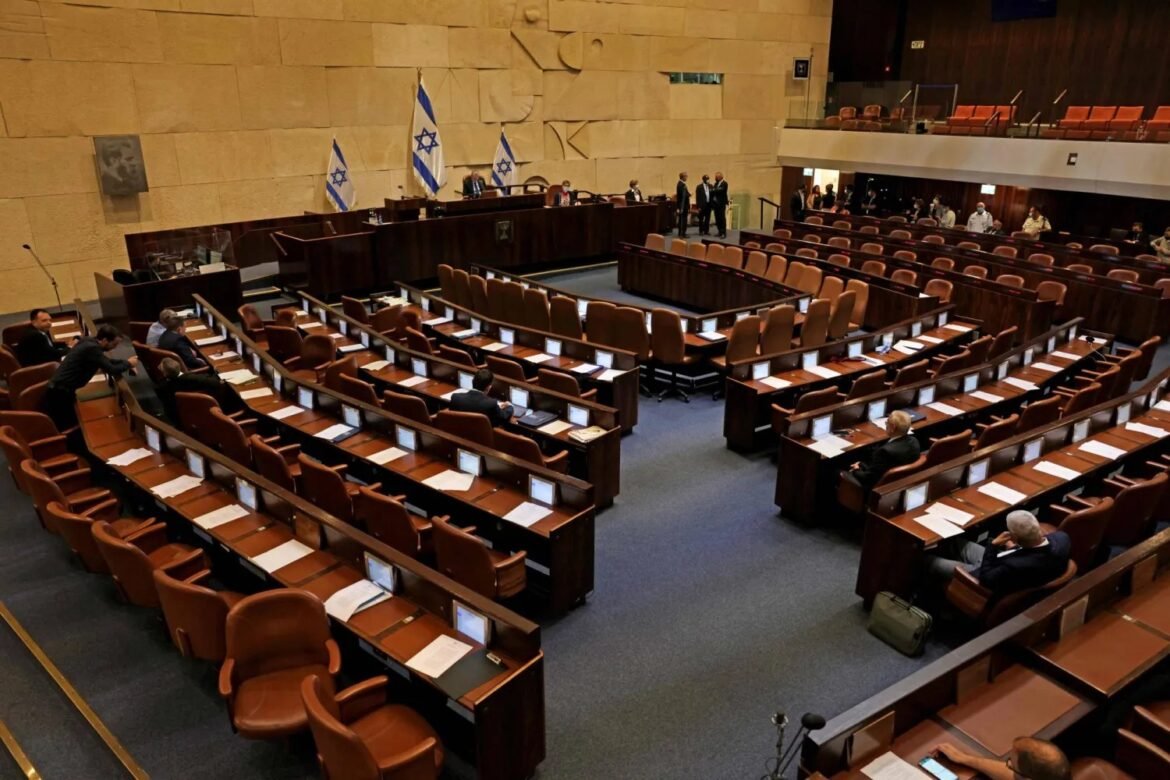 Broadcast-style: A large legislative chamber with curved rows of desks and brown chairs, Israeli flags behind the podium, and a few people seated and standing near the front.
