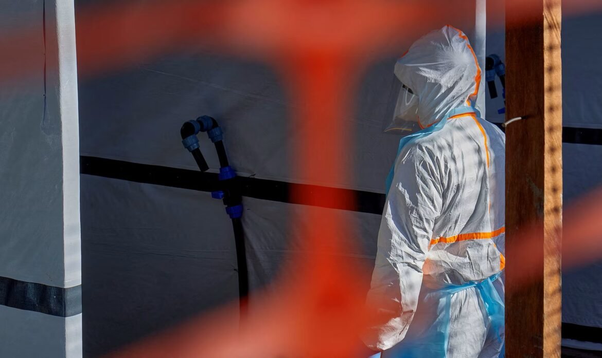 Worker in full white hazmat suit with orange seams stands by a wall with blue pipe fittings, orange barrier in foreground.