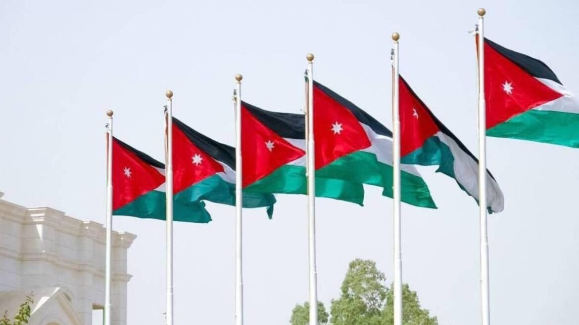 Several Jordanian flags waving on tall flagpoles against a pale blue sky outside a building.