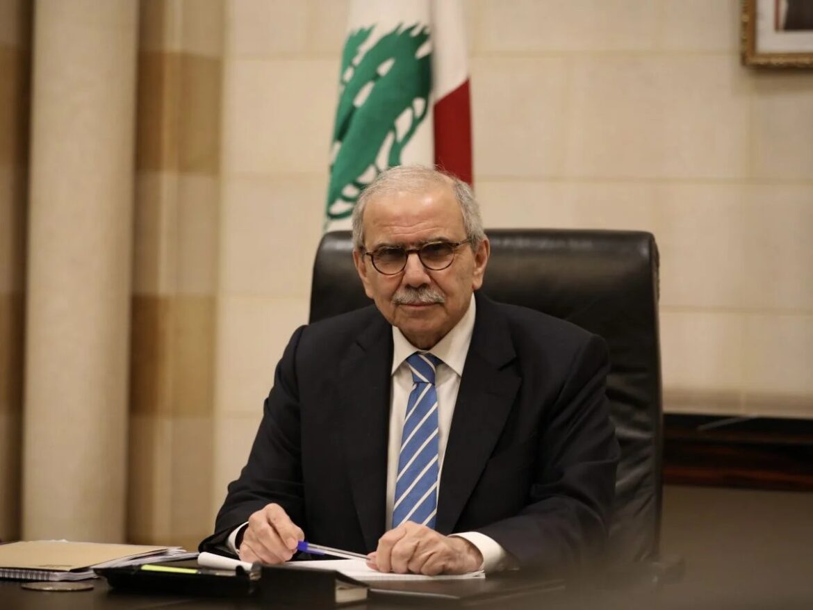 Senior official in a dark suit and glasses sits at a desk, signing papers in an office with a flag in the background.