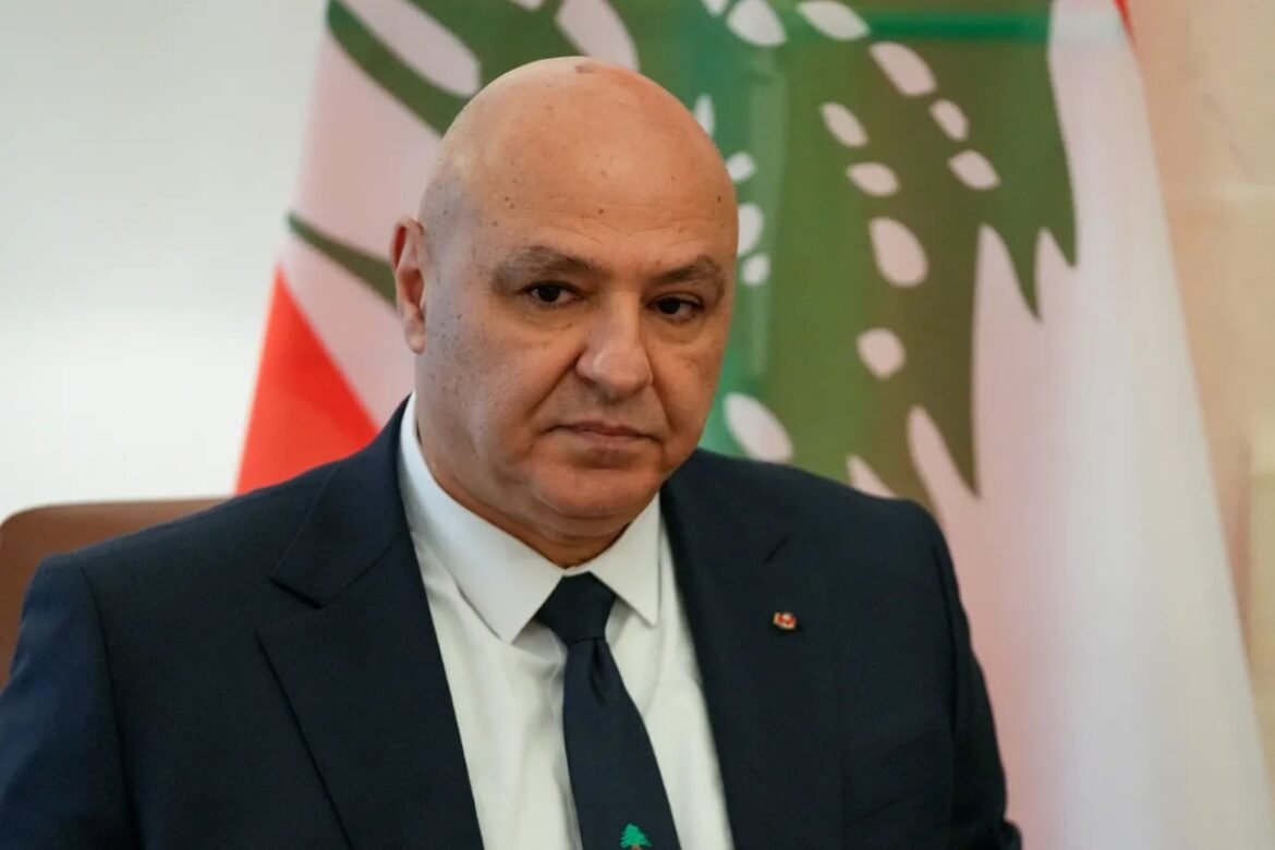 Bald man in a dark suit and tie sitting indoors, with flags in the background, looking serious.