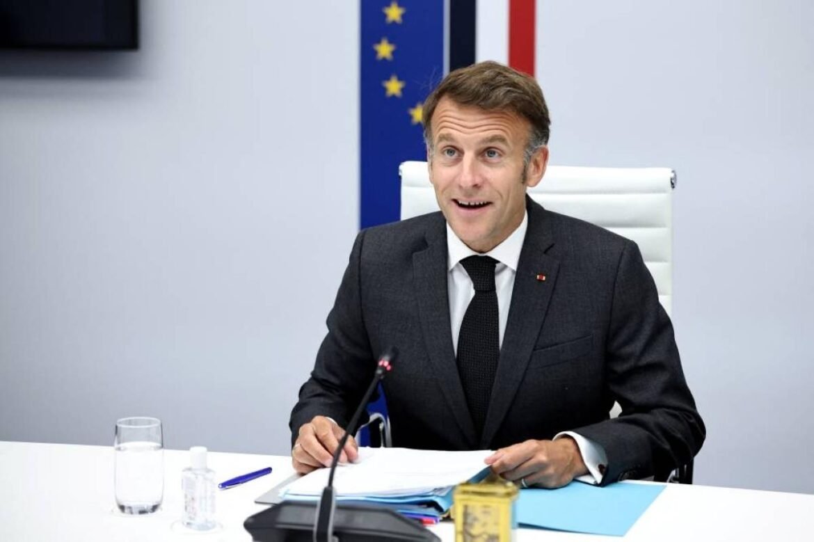 Man in a dark suit and tie sits at a white desk during a formal briefing, speaking into a microphone with papers and a pen in front of him. A glass of water and a small yellow canister sit on the desk, with EU flags in the background.