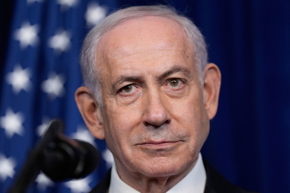Close-up of an older man with gray hair in front of a blue flag backdrop with white stars, facing the camera with a neutral expression.