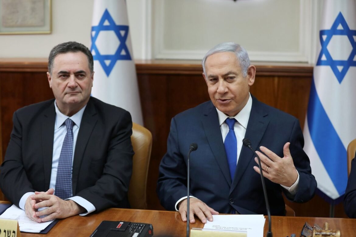 Two men in dark suits sit at a wooden table during a formal meeting, with Israeli flags behind them and microphones in front.