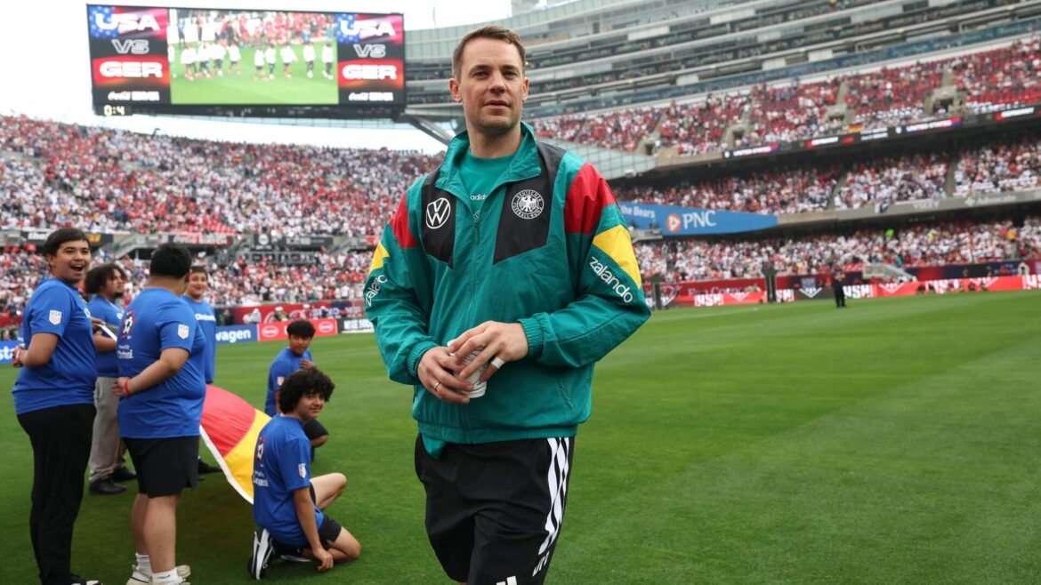 Man in a teal jacket walks toward the camera on a soccer field, with a packed stadium and blue-clad teammates nearby.