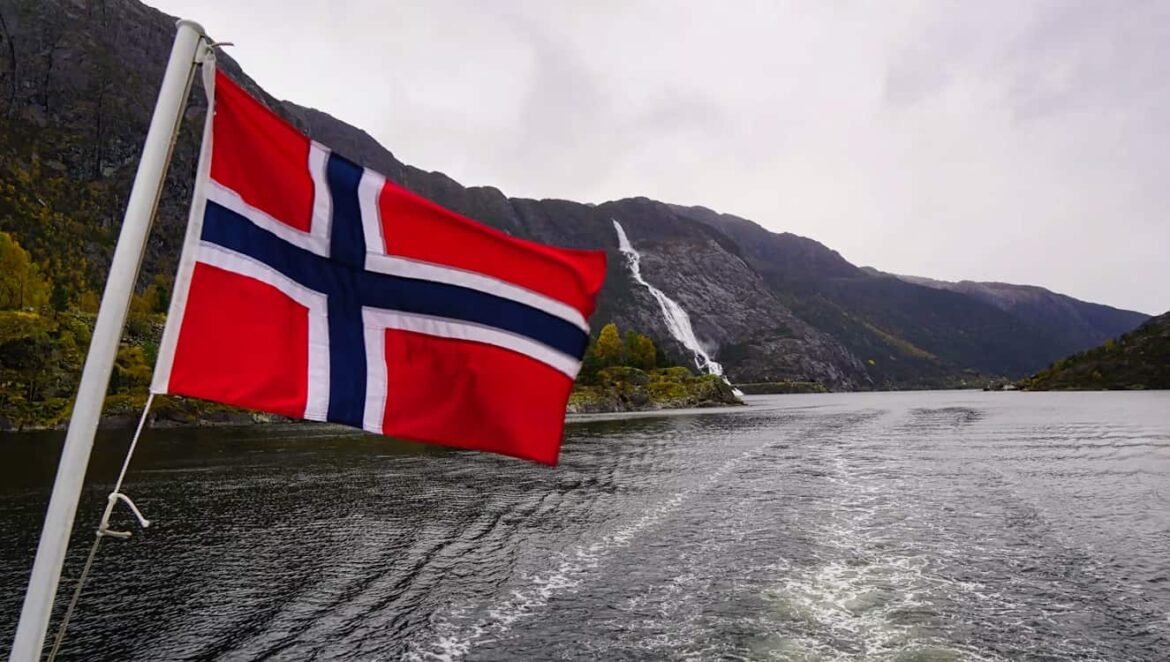 Norwegian flag fluttering from a boat on a fjord with rocky mountains and a waterfall in the background