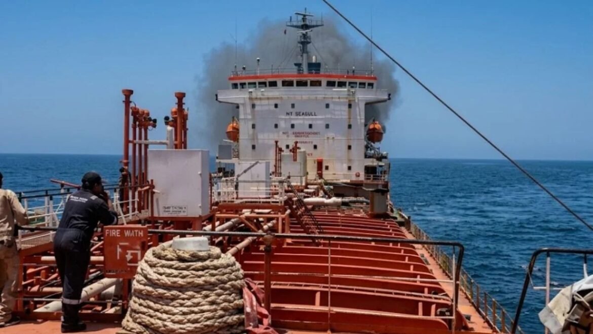 Crew member on the orange oil tanker deck, looking toward a distant white vessel with smoke rising from its stack over the blue sea.