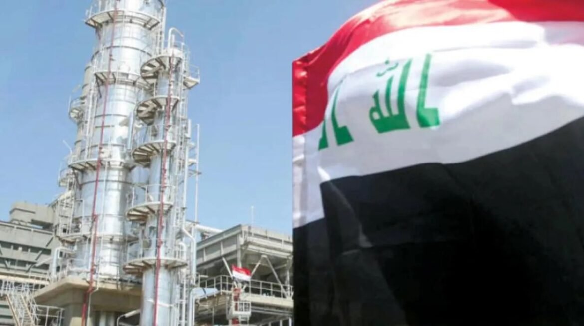 Industrial refinery tower with metal piping beside a waving Iraqi flag against a blue sky, indicating an industrial site in Iraq.