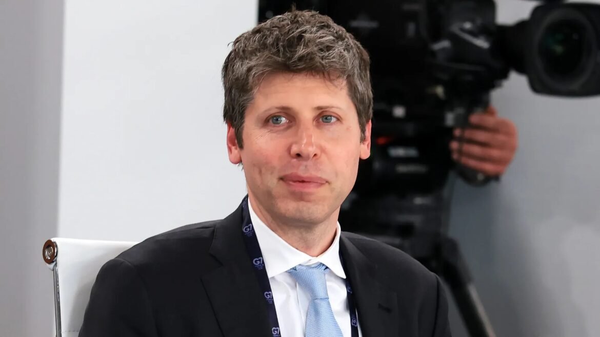 Man in a dark suit and light blue tie sits during an interview, with a camera operator and equipment blurred in the background.