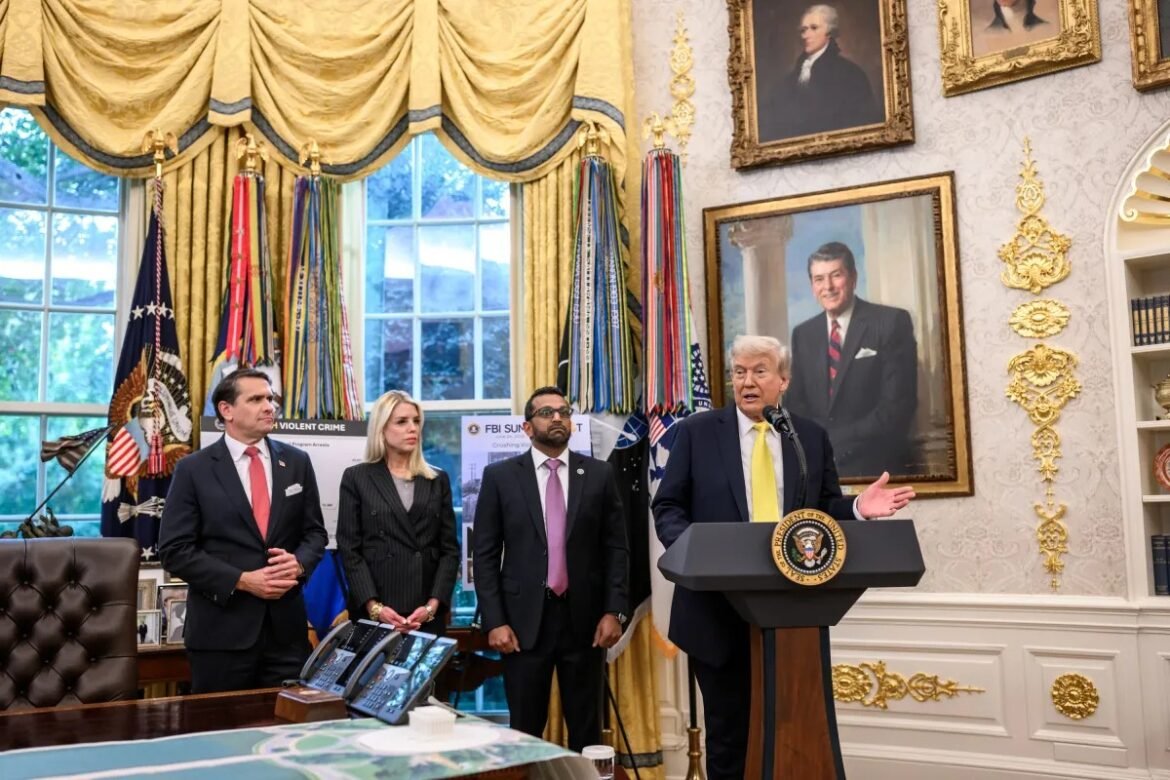 Speaker at a podium giving a briefing in a grand, gold-accented room with flags and portraits behind him, flanked by three aides in formal attire.