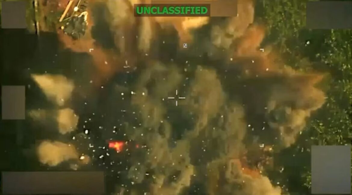 Aerial view of a fiery explosion in a forested area with smoke and flying debris; a center crosshair marks the aiming point, with an UNCLASSIFIED label at the top.