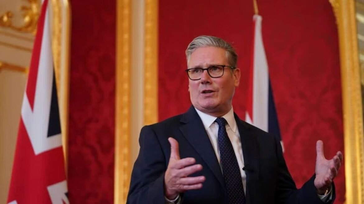 Man in a dark suit and glasses giving a speech in a formal room with red wallpaper and flag behind him.