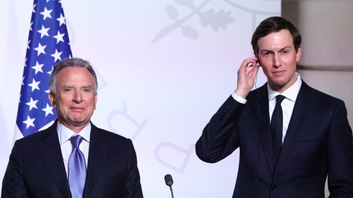 Two men in dark suits stand at a podium in front of an American flag during a formal event.